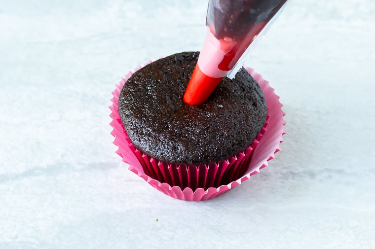 Raspberry jam being piped into the center of a chocolate cupcake over a white background