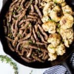 A cast iron skillet with garlic butter steak bites on one side and cauliflower florets on the other with a blue and white striped napkin and fresh thyme sprigs all around over a white background