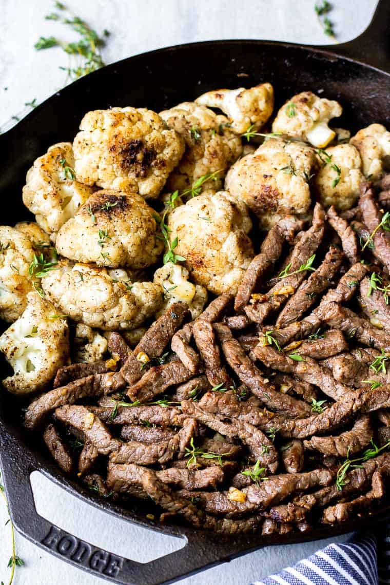 Close up of a cast iron skillet with garlic butter steak bites on one side and cauliflower florets on the other with a few pieces of fresh thyme in the background over a white backdrop