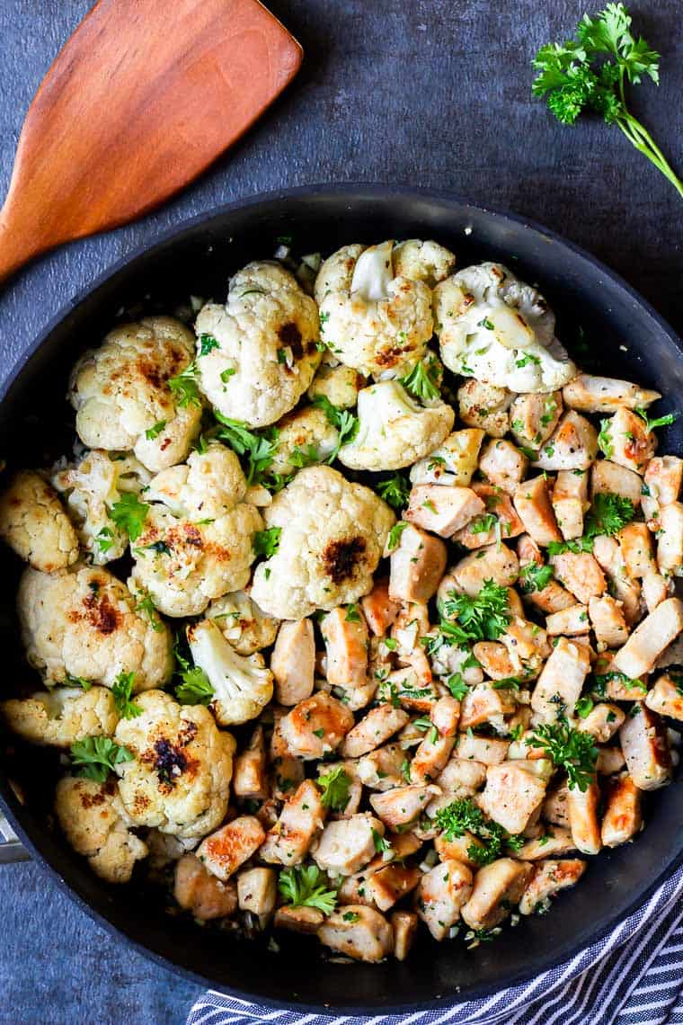 Garlic Butter Chicken and Cauliflower in a black skillet with a wood spatula, blue and white striped towel, and fresh parsley around it on a blue gray background