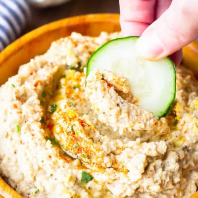 A slice of cucumber being dipped into a wooden bowl of cauliflower hummus with a bulb of garlic, a lemon, and a part of a striped towel in the background