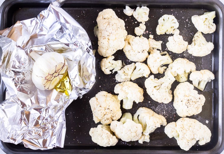 Raw cauliflower florets and a bulb of garlic on foil on a baking sheet before roasting