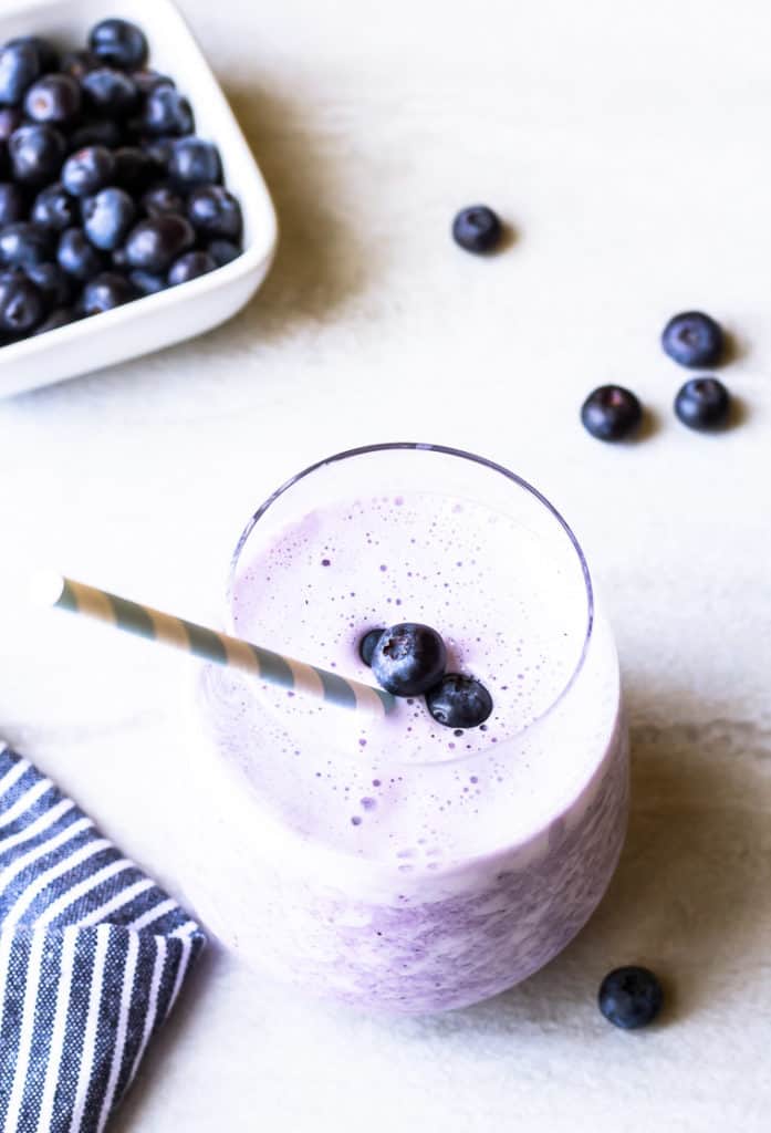 A keto blueberry smoothie in a glass cup with a blue and white paper straw and part of a blue and white striped towel, a square white bowl full of blueberries, and extra blueberries in the background on a white backdrop