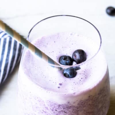 A keto blueberry smoothie in a glass cup with a blue and white paper straw and part of a blue and white striped towel and extra blueberries in the background on a white backdrop