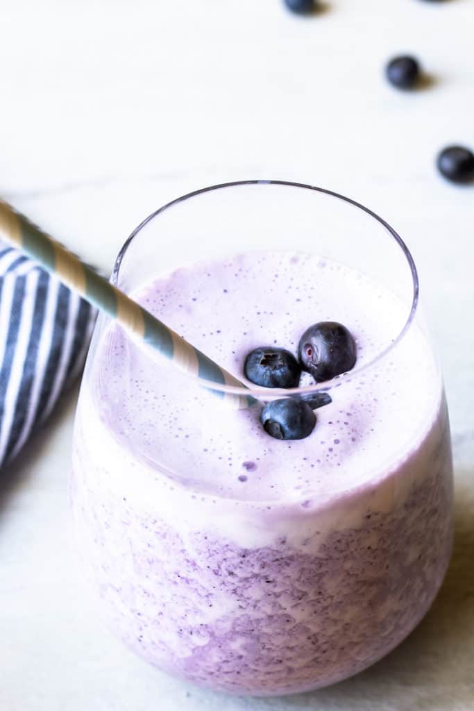 A keto blueberry smoothie in a glass cup with a blue and white paper straw and part of a blue and white striped towel and extra blueberries in the background on a white backdrop