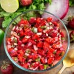 Strawberry salsa in a clear bowl with extra limes, cilantro and strawberries around it