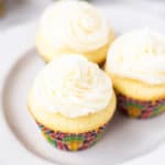 3 mini vanilla cupcakes on a small white plate with 3 more blurred in the background on a white backdrop