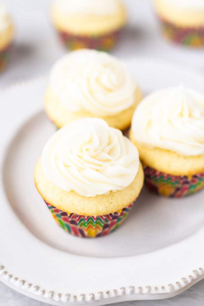 3 mini vanilla cupcakes on a small white plate with 3 more blurred in the background on a white backdrop