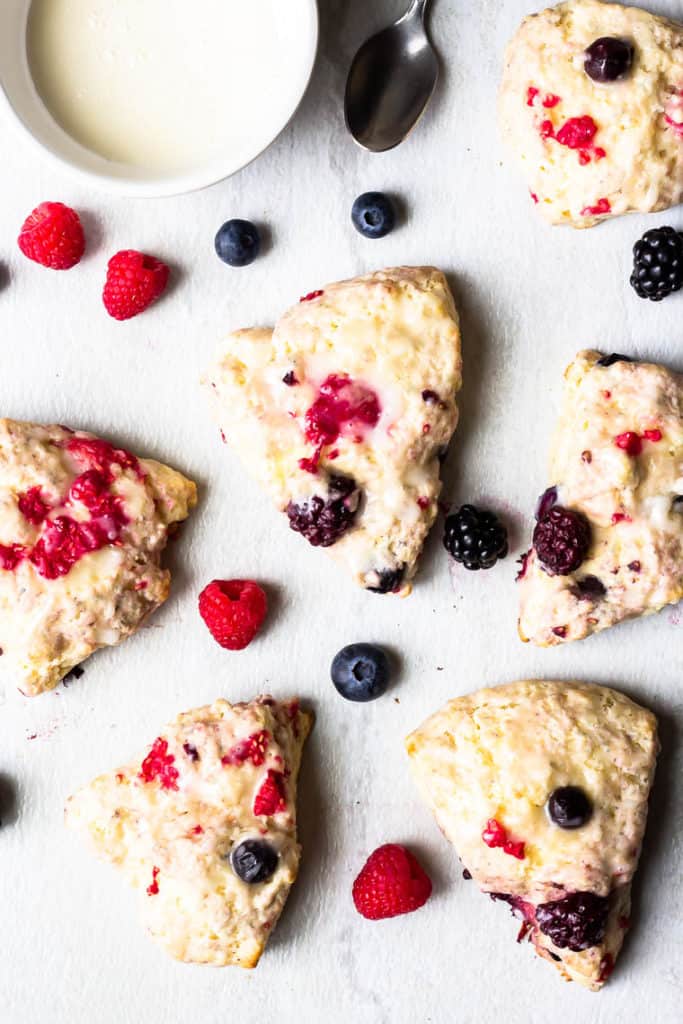 Overhead of 6 berry scones on a white background with fresh berries scattered around them, a small white bowl of glaze and a spoon