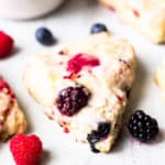 Close up of a mixed berry scone on a white table with fresh berries, parts f 2 more scones, and a small white bowl all around it