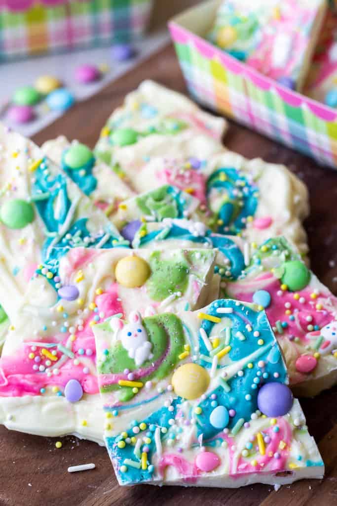 White chocolate easter bark broke into pieces on a wood board with easter boxes filled with more bark and loose candy and sprinkles in the background