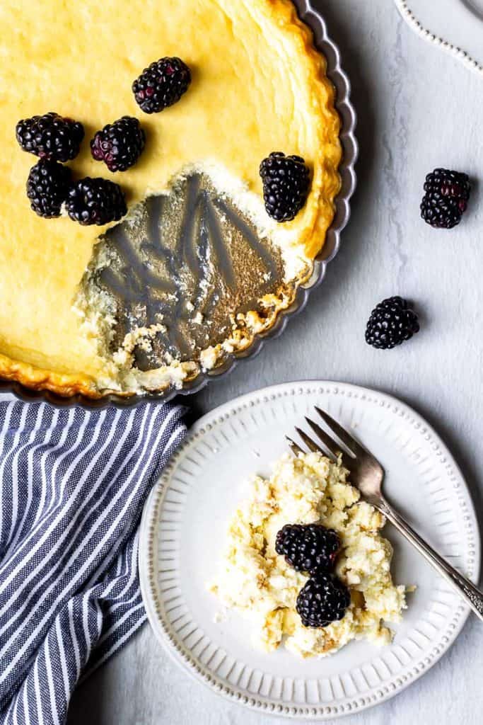 Cheesecake in a tart pan with a serving spooned out onto a white plate with a fork and topped with blackberries. There is a blue and white striped towel between the pan and plate and 2 blackberries above the plate all over a white background