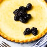 Close up of a crustless baked cheesecake topped with blackberries with a fork and part of a blue and white striped napkin in the foreground over a white background
