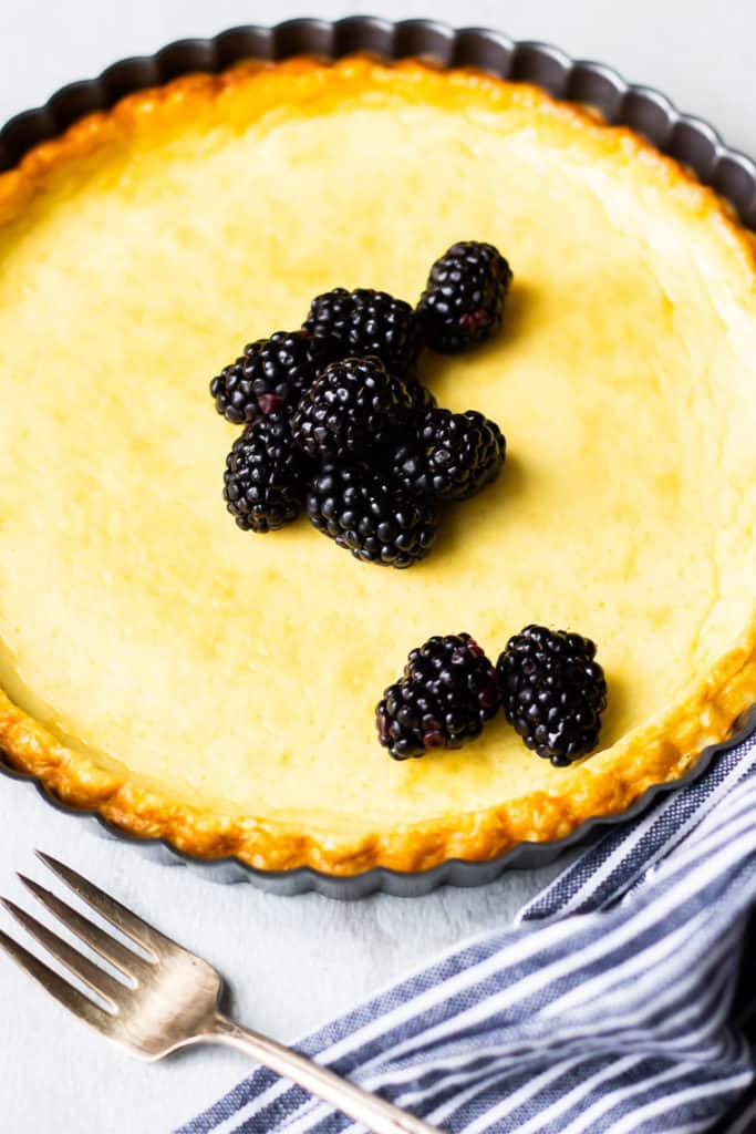 Close up of a crustless baked cheesecake topped with blackberries with a fork and part of a blue and white striped napkin in the foreground over a white background