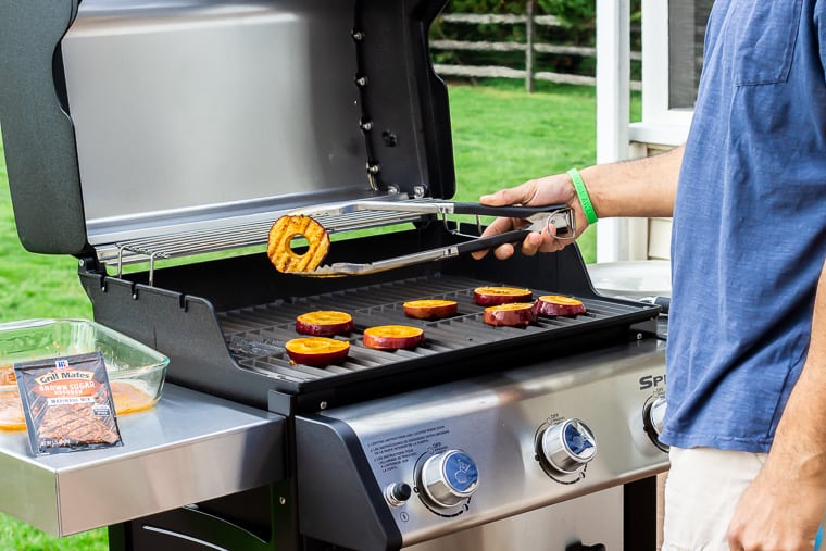 A person holding up a grilled apple slice over the grill with more slices still cooking on it. There is grass, a part of a wood fence, and a packet of seasoning in the background