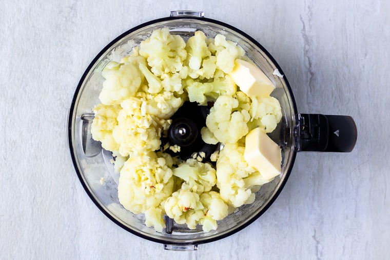 Steamed cauliflower, garlic and butter in a food processor bowl with a blade over a white background
