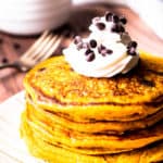 Close up of a stack of pumpkin chocolate chip pancakes topped with whipped cream and mini chocolate chips with a fork and white bowl full of chips faded in the background all over a wood background
