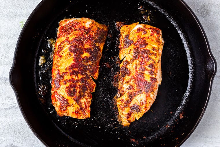2 blackened cod fillets in a black cast iron pan over a white background