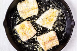 4 Cod fillets in lemon garlic butter sauce in a cast iron skillet over a white background