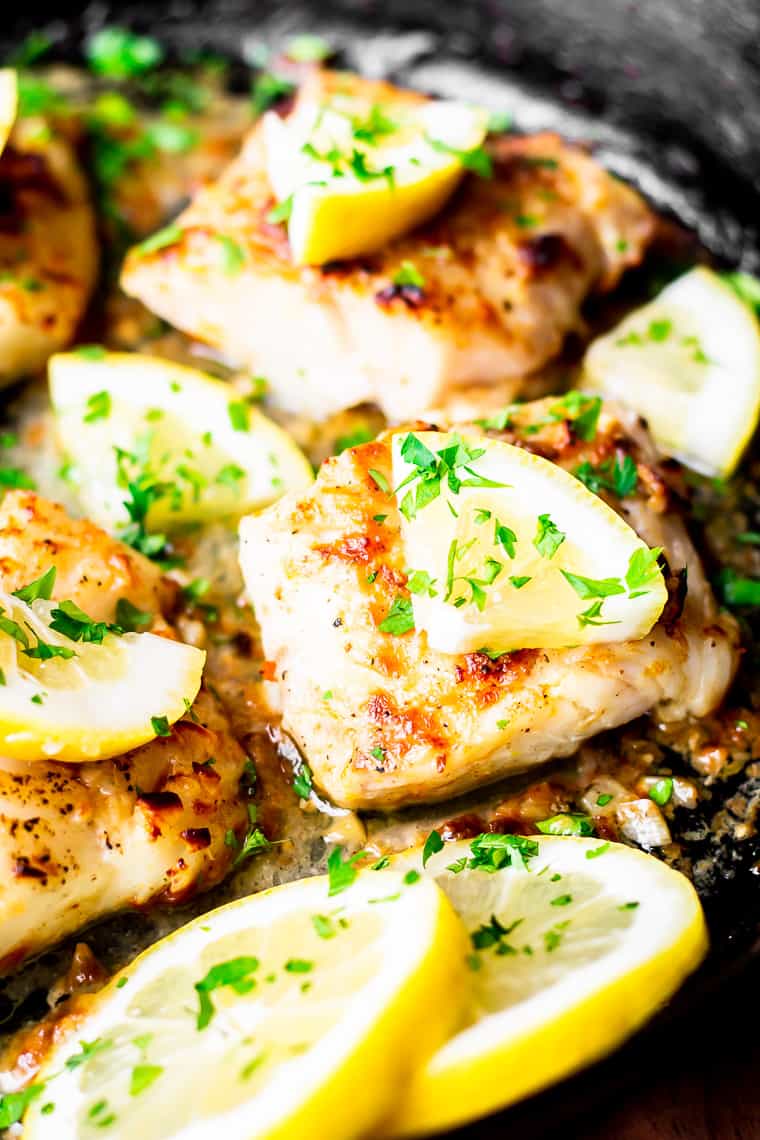 Close up of 2 cod fillets in a cast iron skillet with pieces of lemon, lemon slices, and parsley on them.