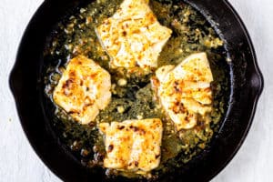Broiled cod fillets in a lemon garlic butter sauce in a cast iron skillet over a white background