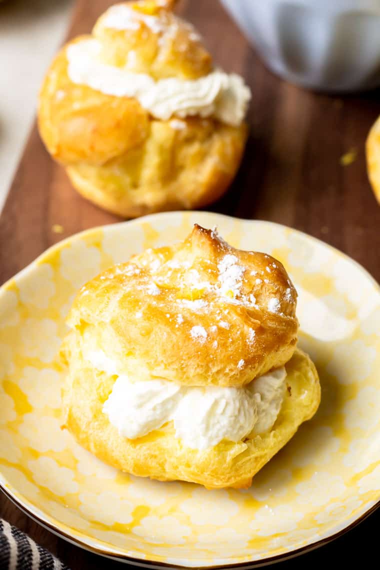 One lemon cream puff on a small plate over a wood table with a second puff and small blue bowl in the background