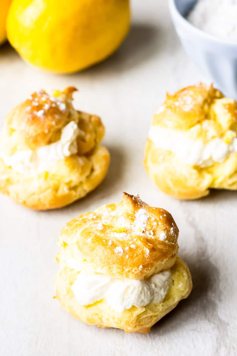 3 lemon cream puffs on a white background with lemons and small bowl behind them