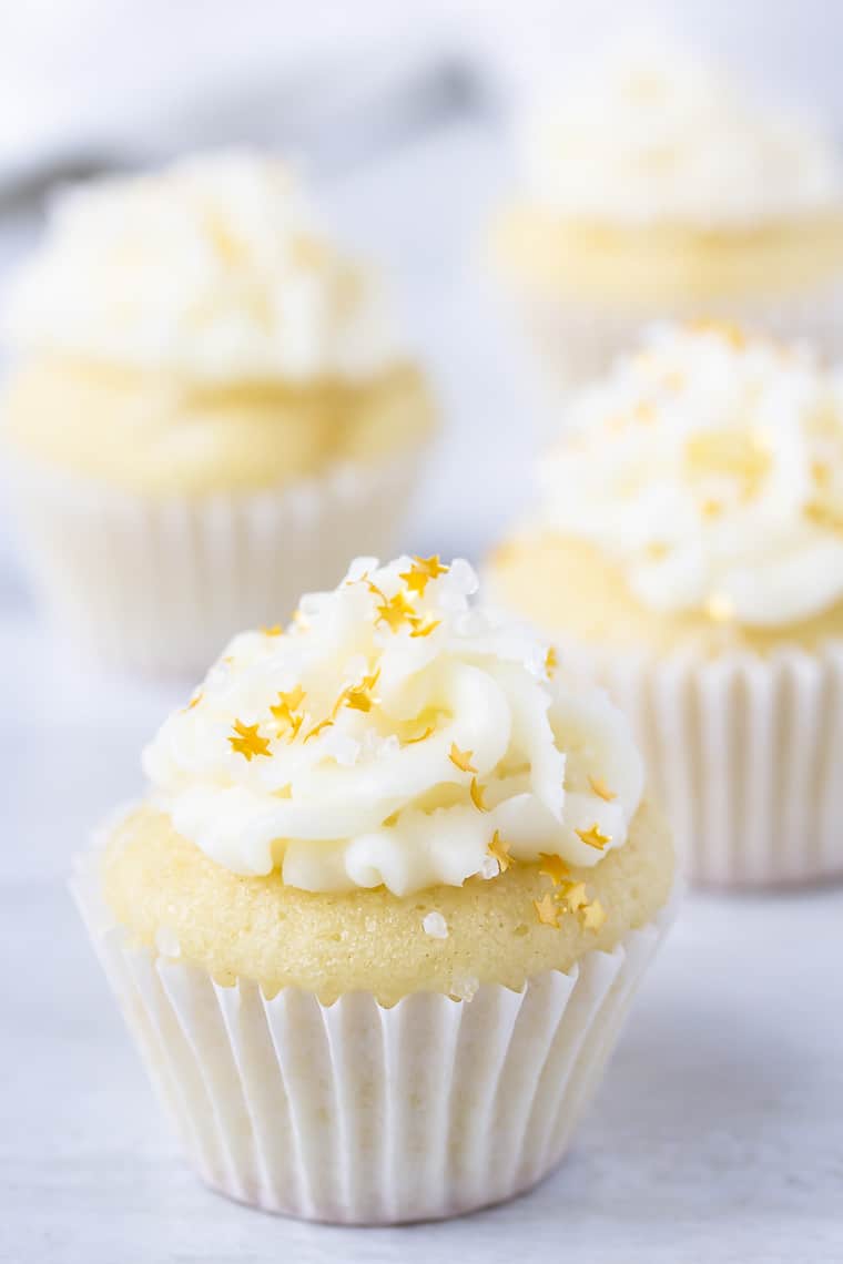 Close up of a mini coconut cupcakes topped with sugar and gold stars on a white background with additional cupcakes blurred in the background