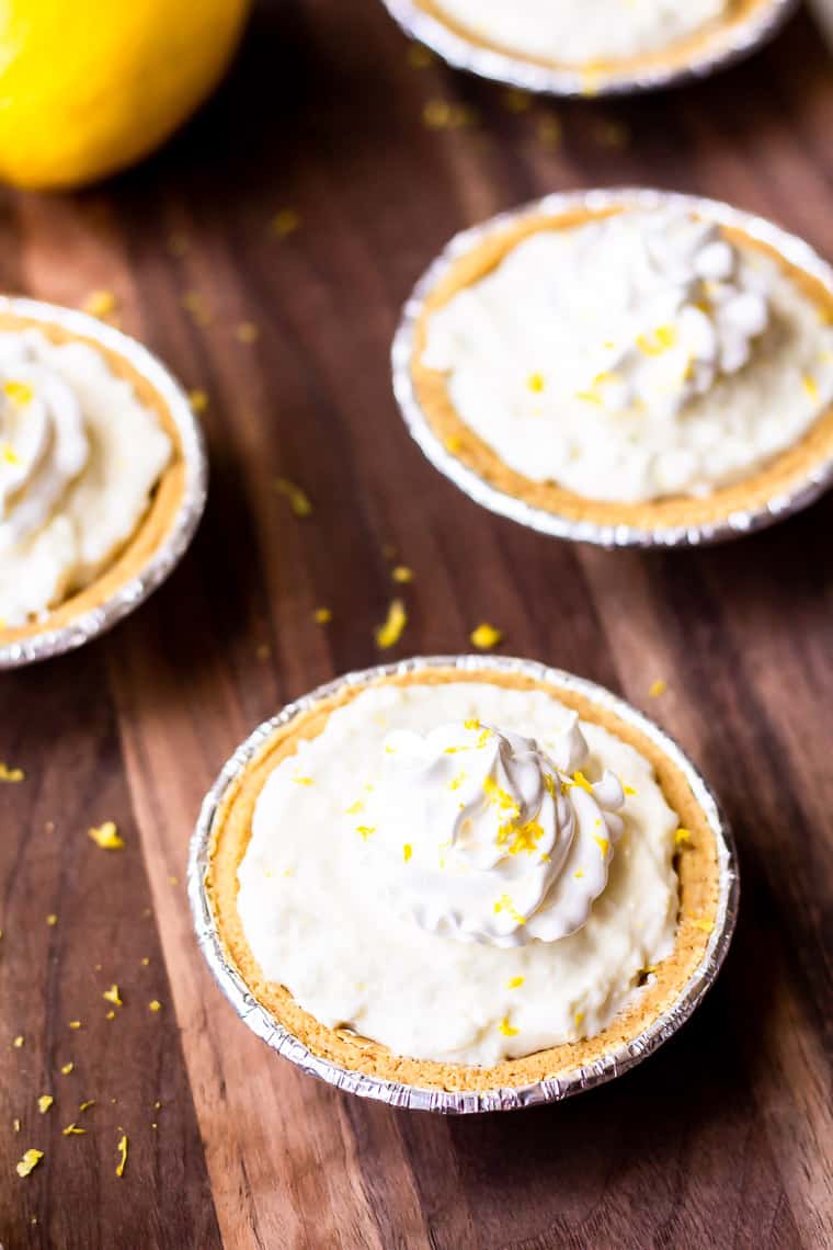 4 mini lemon cheesecakes with a lemon and lemon zest around them on a wood background