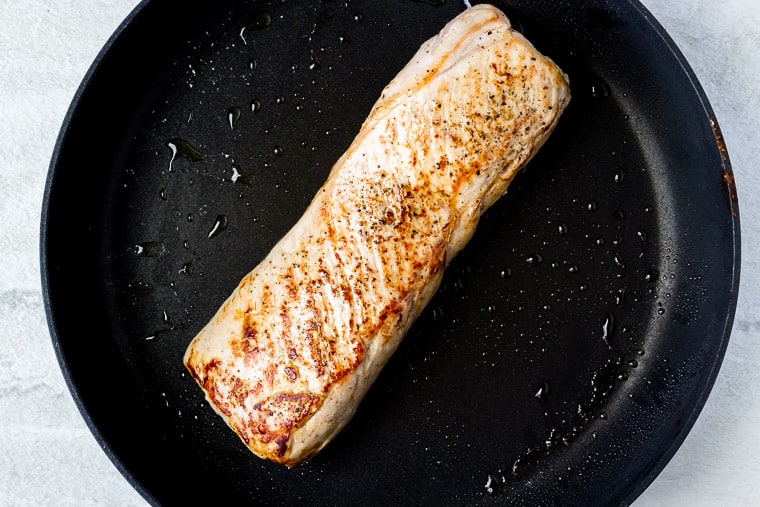 Browned pork loin in a black skillet over a white background
