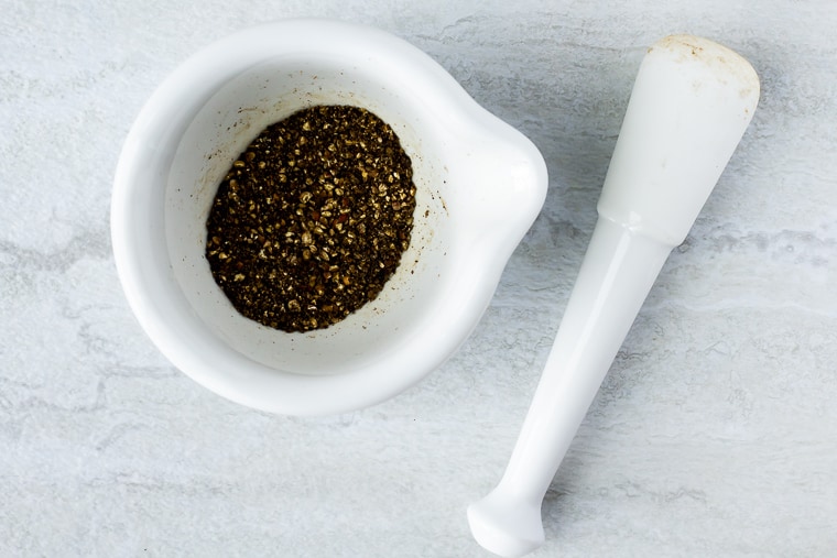 Black peppercorns in the bowl of a mortar and pestle with the pestle sitting next to it on a white background