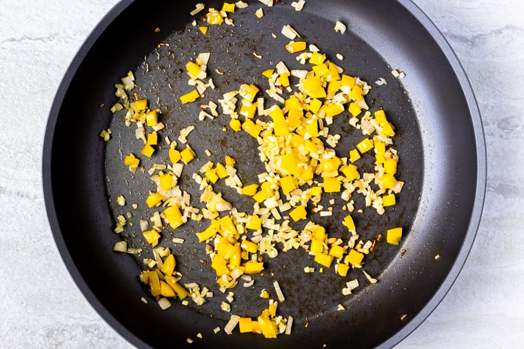 Yellow pepper, shallot, and garlic cooking in a black skillet over a white background