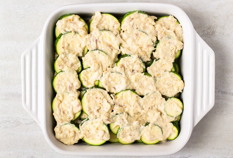Zucchini rounds layered in a white casserole dish and topped with cheese sauce on a white backdrop