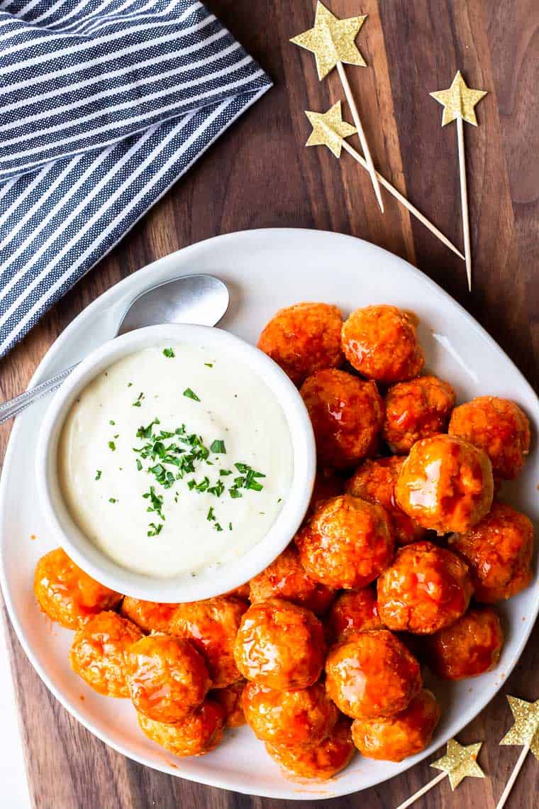 Buffalo chicken meatballs on a white plate with a bowl of ranch dressing on a wood board with a blue and white striped towel and star toothpicks