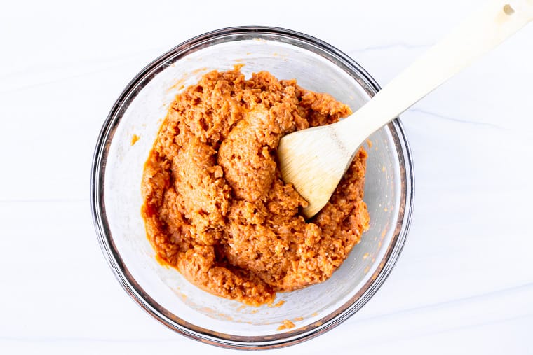 Keto buffalo chicken meatball mixture in a glass bowl with a wood spoon over a white background