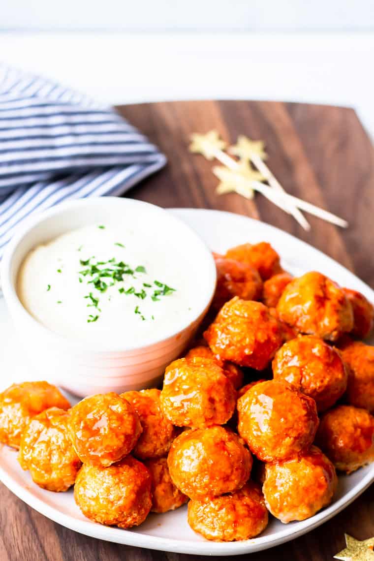 Keto buffalo chicken meatballs on a white plate with a bowl of ranch dressing on a wood board with a blue and white striped towel with a white background