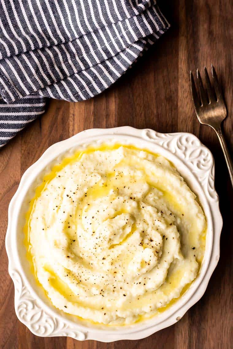 Overhead of creamy mashed cauliflower with butter melted on it on a wood table with a fork and blue striped towel