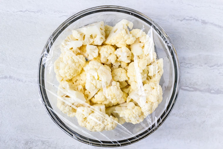 Cauliflower florets in a glass bowl covered with plastic wrap on a white background