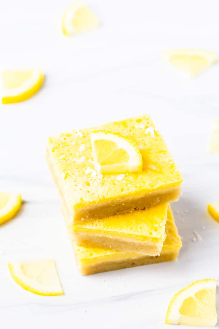 3 keto lemon bars stacked on top of each other with a lemon slice on top and a few more around them