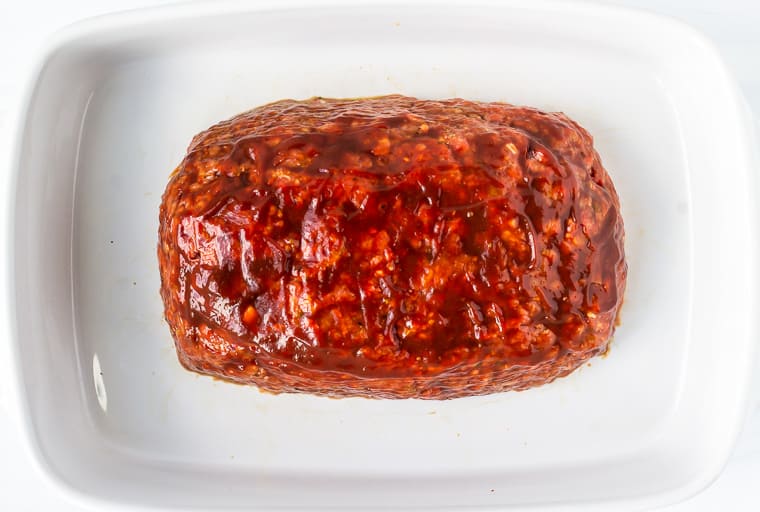 Raw meatloaf basted with barbecue sauce in a white baking dish