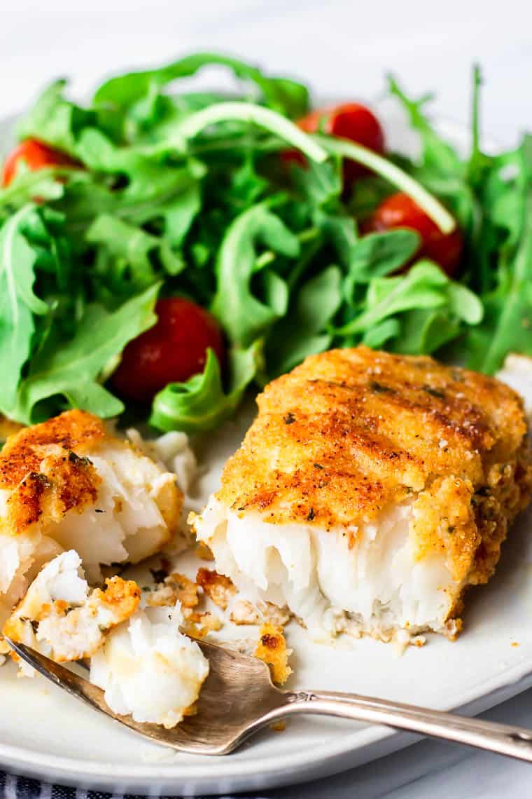 Pan fried cod on a plate cut in half with a fork and a salad