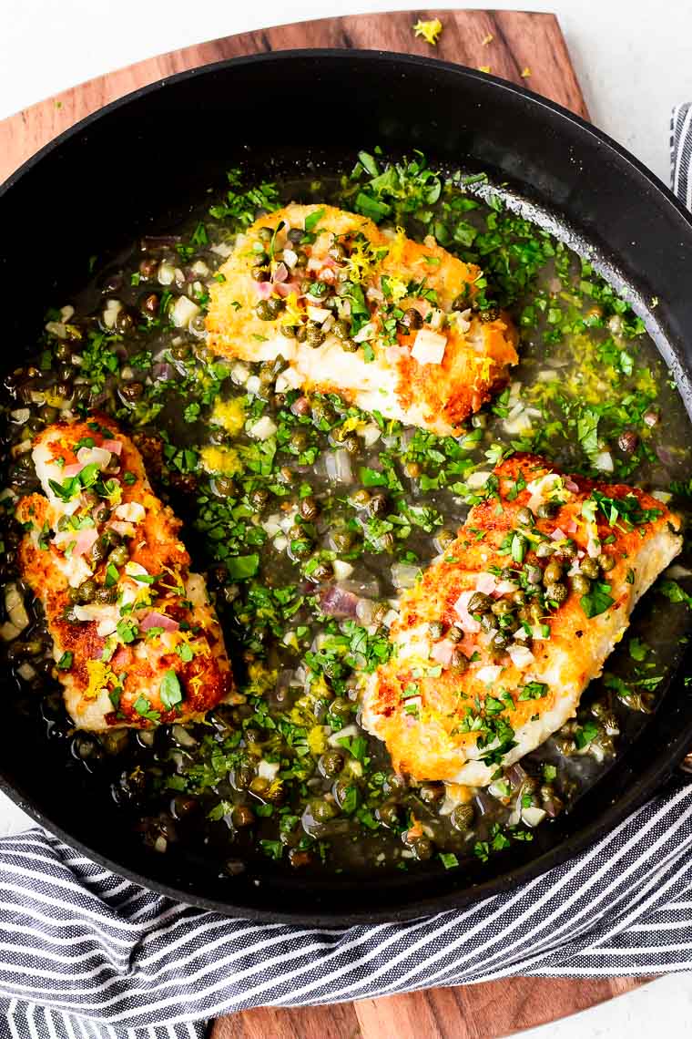 3 cod fillet in piccata sauce in a black skillet over a wood board with a blue and white striped towel around the bottom edge of the pan