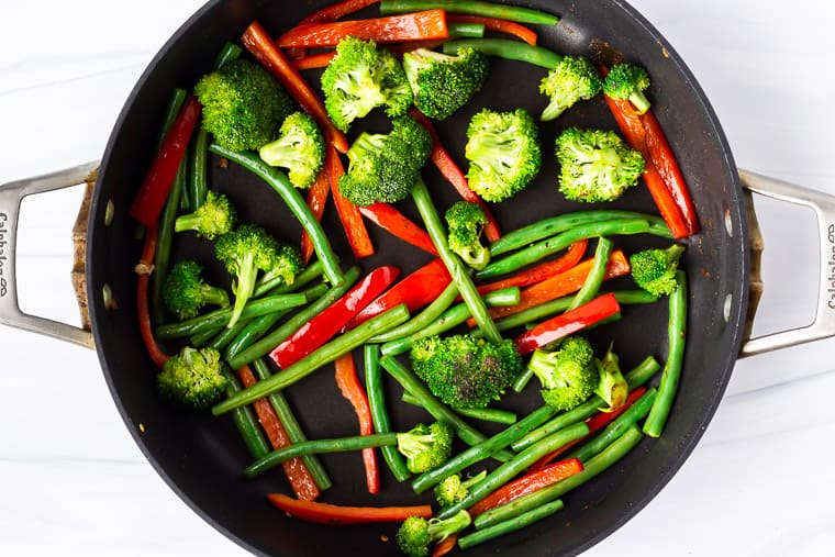 Broccoli, green beans, and peppers cooking in a black skillet over a white background