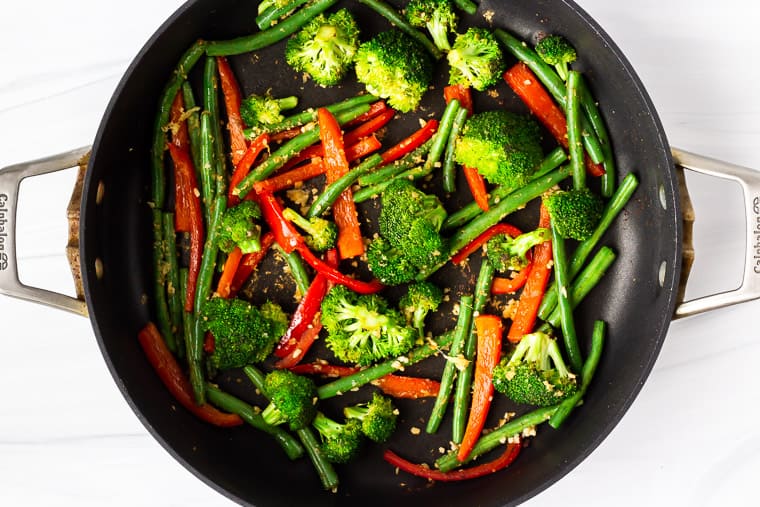 Broccoli, green beans and peppers cooking with garlic and ginger in a black skillet over a white background