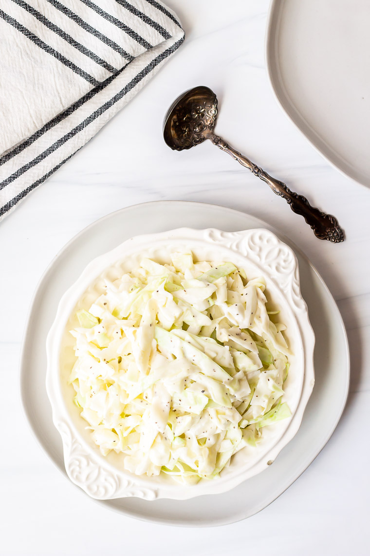 Overhead of keto coleslaw in a white bowl on a white plate on a white background with an antique spoon, black striped white towel and a second white plate