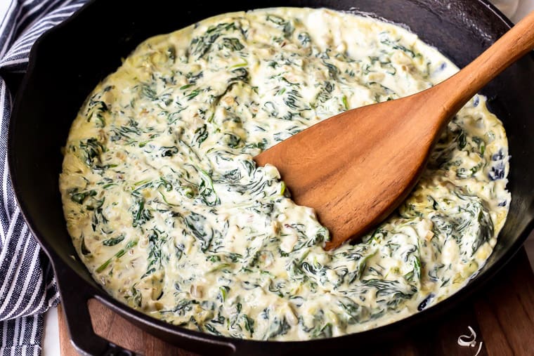 Keto Creamed Spinach in a black cast iron pan with a wood turner in it and a wood board underneath with a blue and white striped towel behind the pan