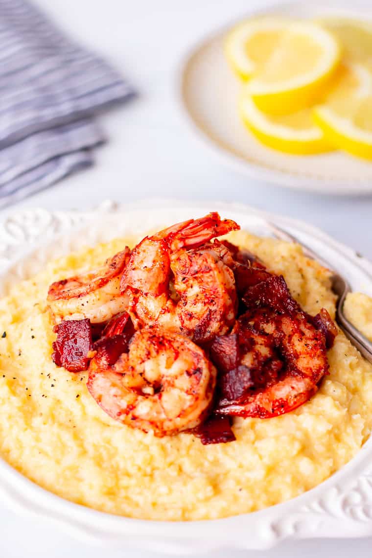 Keto Cajun Shrimp and Cauliflower Grits in a white bowl on a white background with a white plate of lemon slices and a blue and white towel in the background
