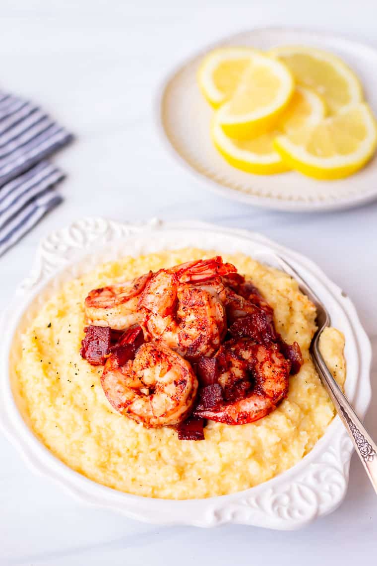 Keto Cajun Shrimp and Grits in a white bowl on a white background with a white plate of lemon slices and a blue and white towel in the background