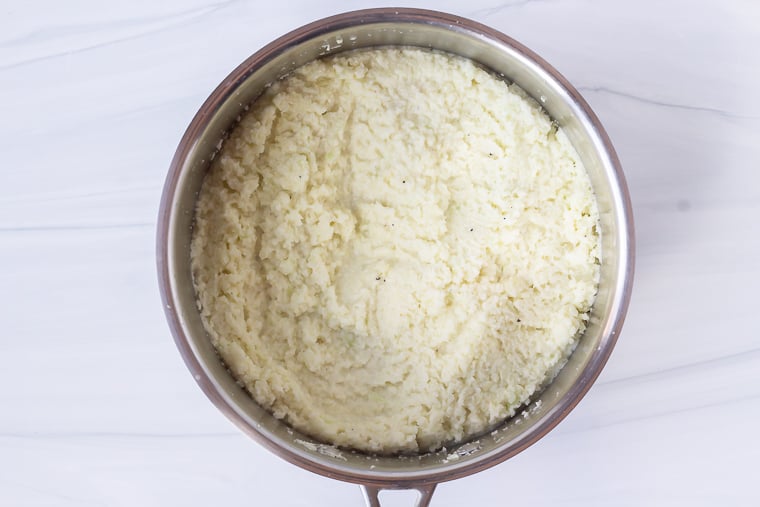 Cauliflower grits cooking in a silver pot over a white background