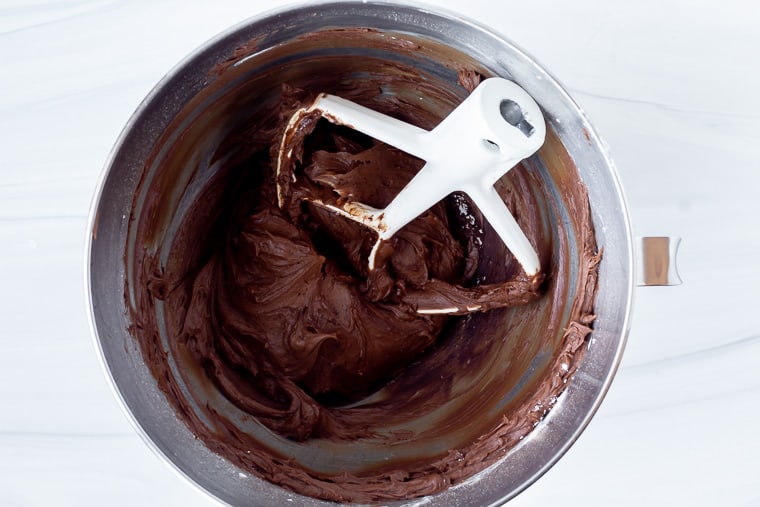 Chocolate frosting in a silver mixing bowl with the mixing paddle in it to show thickness